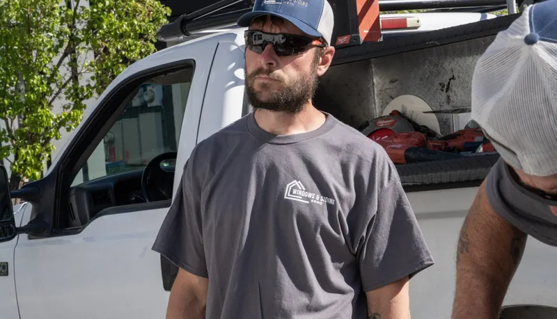 A bearded Windows and Siding Reno installer in sunglasses and a logo tee stands in front of a white work truck
