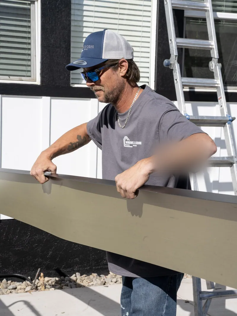 One of our crew carrying a long formed fascia piece along the side of a house with an extension ladder behind him