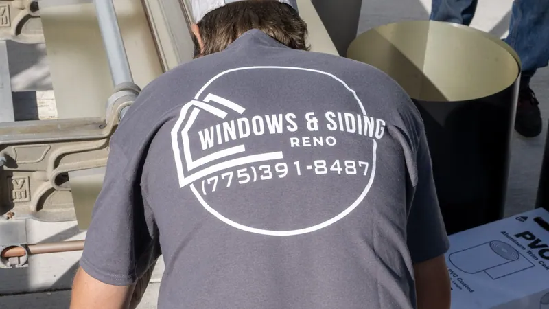 A Windows and Siding Reno installer, logo and phone number on his shirt back, bends over a brake beside a trim coil roll