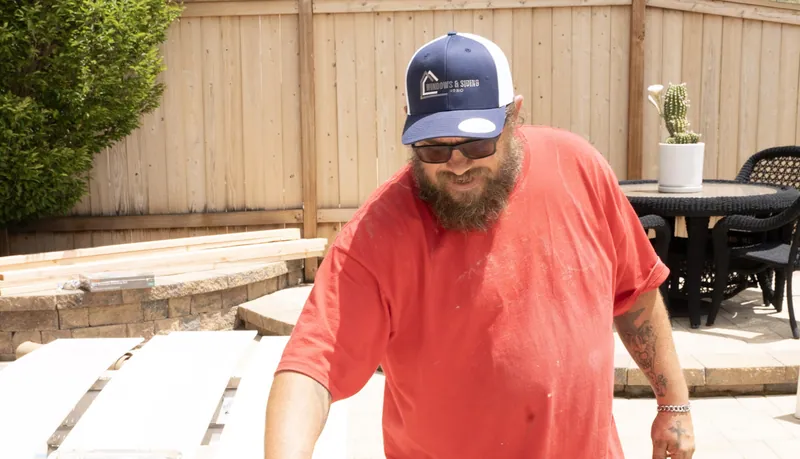 A bearded Windows and Siding Reno crew member in the logo cap stands over long white panels laid out on a backyard patio