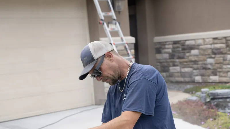 A crew member in a cap and sunglasses leans forward in a driveway, a garage door, ladder and stone veneer behind him