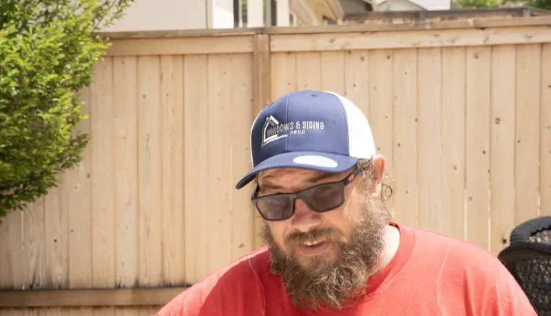 A bearded Windows and Siding Reno crew member in the logo cap and sunglasses talks in a backyard by a wood fence