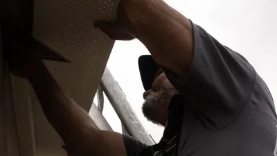 A crew member holds a perforated soffit panel up under an eave and checks it against a speed square in his other hand