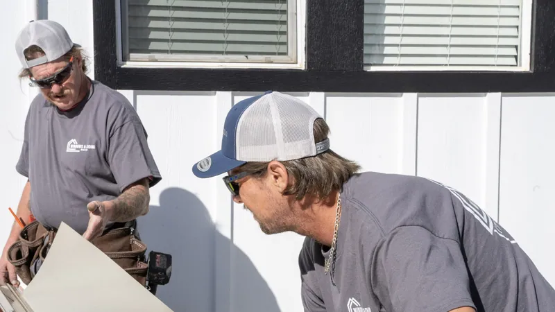 Two installers in company shirts handling a formed trim sheet beside a white board-and-batten wall