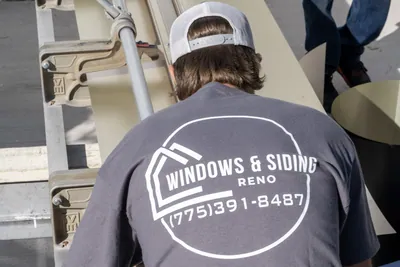 A Windows and Siding Reno installer, logo and phone number on his back, bends over tan trim coil on a portable brake