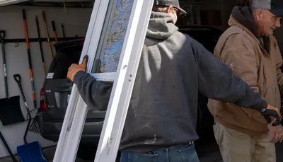 An installer carries a tall new white vinyl window past an open garage as an older coworker in a tan jacket stands at right