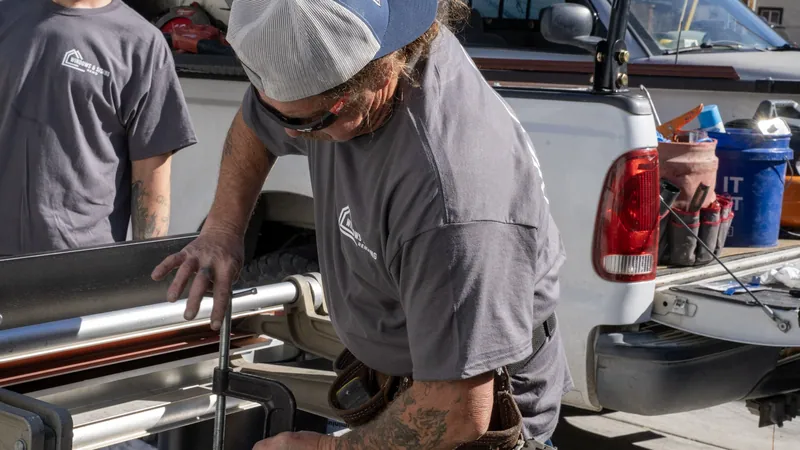 A Windows and Siding Reno installer tightens a C clamp on a sheet metal brake loaded with trim coil beside a white pickup