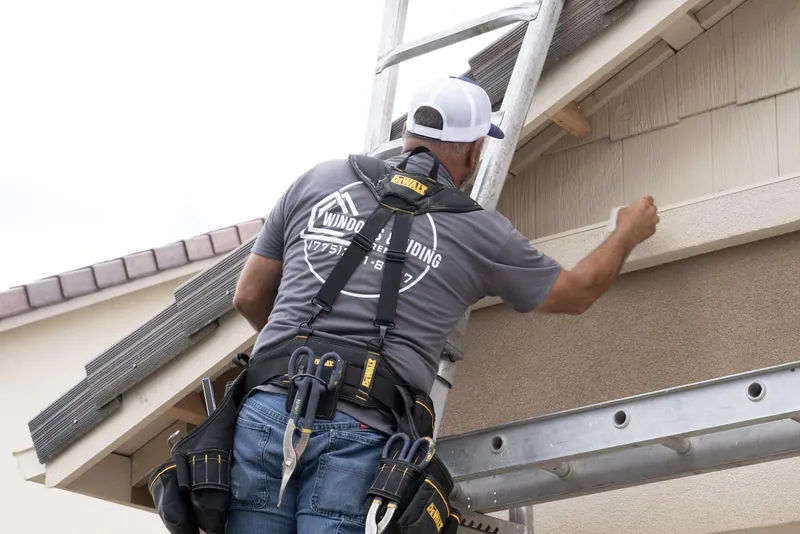 A Windows and Siding Reno installer on a ladder works along the joint where gable siding meets a stucco band