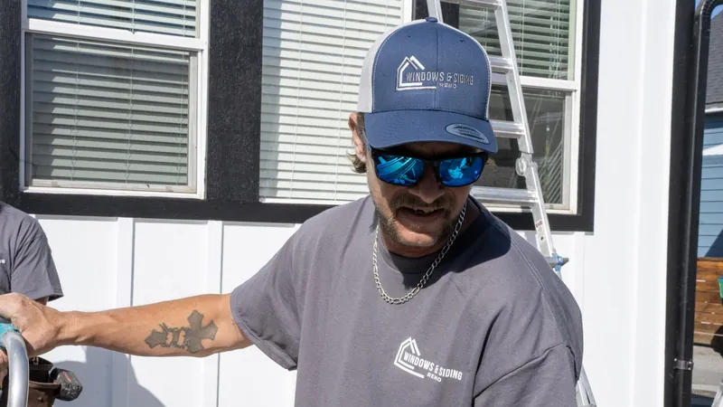 A Windows and Siding Reno installer in a logo cap grips an aluminum rail below white windows with black trim on white siding
