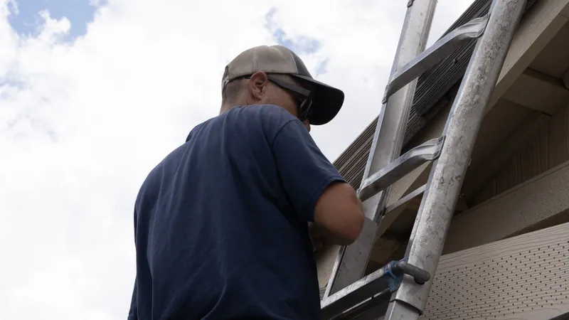 An installer in a cap and safety glasses works from an aluminum ladder at the eave beside perforated soffit panels