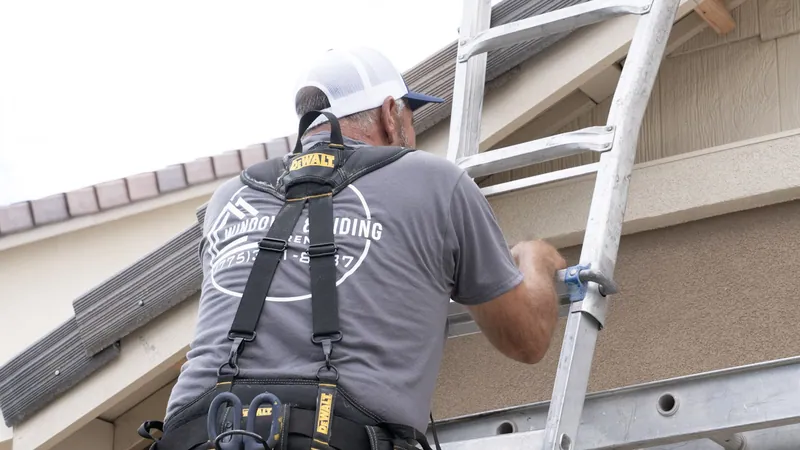 A Windows and Siding Reno installer steadies a ladder rig below a gable with shake style siding under a tile roof