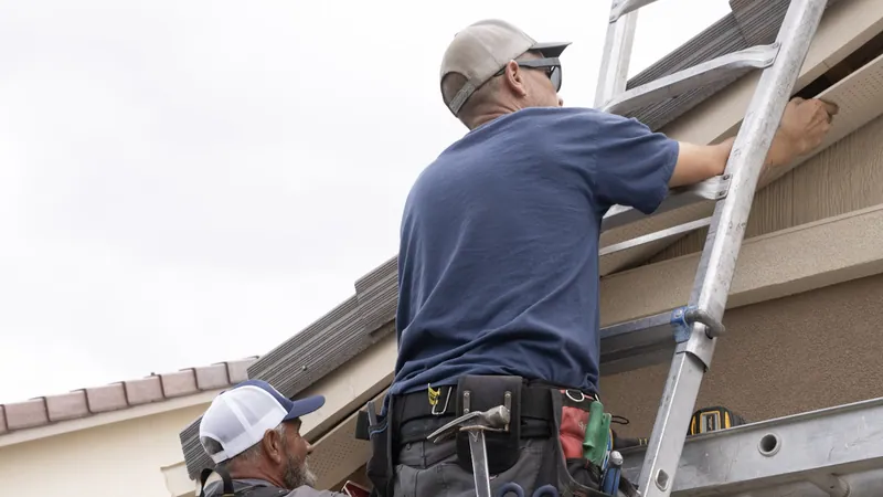 An installer on an aluminum extension ladder presses a perforated soffit panel under a tile roof eave, a coworker below