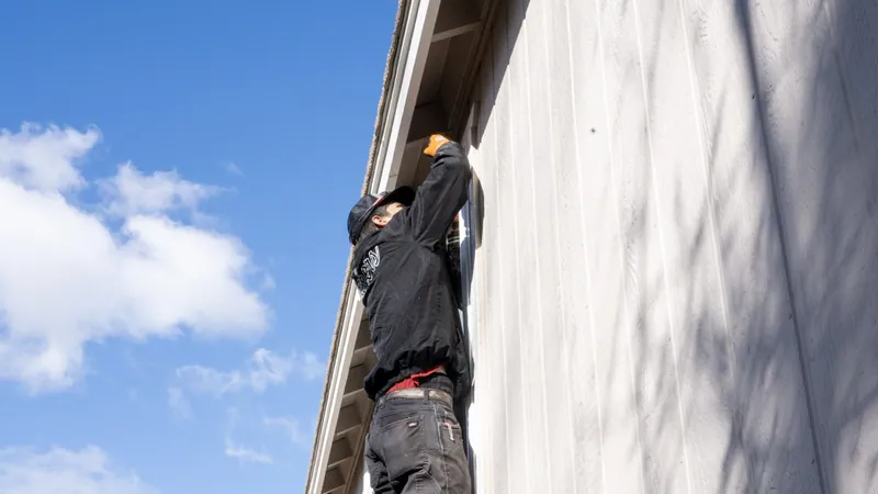 An installer in a black jacket reaches up to the top of a window just under the eave of a house with light vertical siding