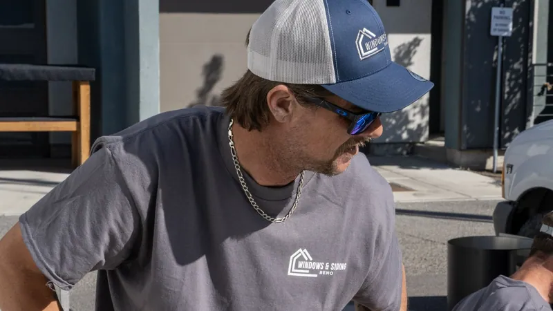 A Windows and Siding Reno installer in a logo cap, sunglasses and logo tee leans forward and looks down on a sunny street