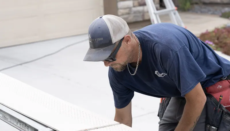 A Windows and Siding Reno installer in a navy logo tee lines up white perforated soffit panels on a saw rail in a driveway