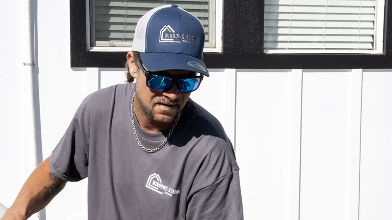 A Windows and Siding Reno installer in a logo cap and logo tee looks down below black trimmed windows on white siding