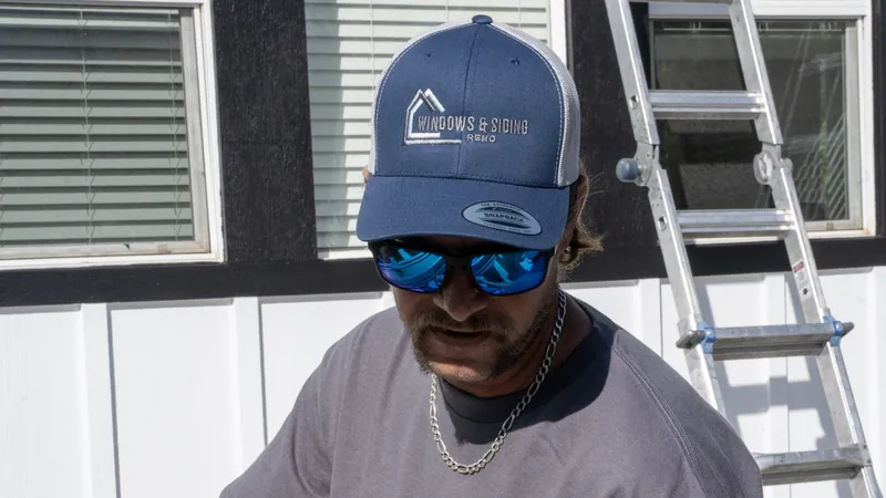A Windows and Siding Reno installer in a logo cap and mirrored sunglasses looks down by white windows set in black trim