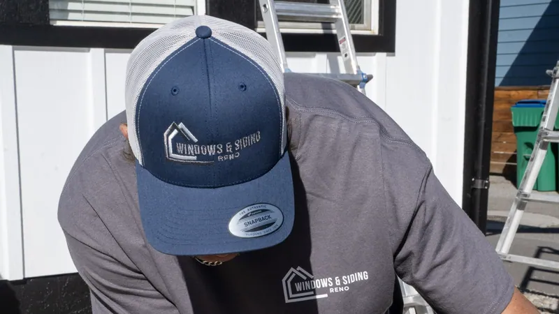 A Windows and Siding Reno installer in a logo cap and tee bends over his work beside white board and batten siding