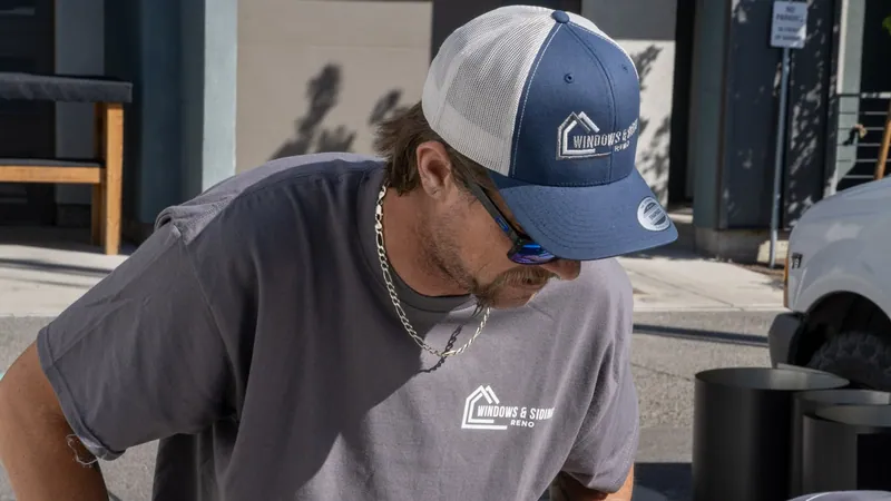 A Windows and Siding Reno installer in a logo cap and tee leans forward beside black coil rolls and a white truck