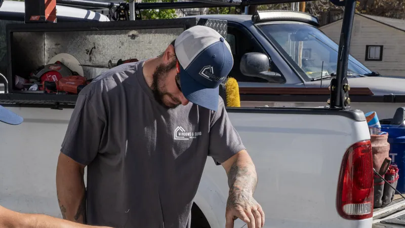 A Windows and Siding Reno installer in the logo cap and tee looks down at his work beside a pickup loaded with tools