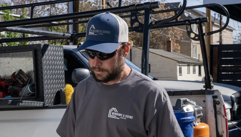 A Windows and Siding Reno installer in a logo cap and tee stands beside a white work pickup with ladder rack and toolbox