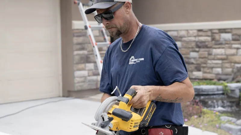 A Windows and Siding Reno installer in a navy logo tee holds a yellow cordless circular saw in front of a garage