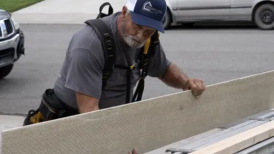 A Windows and Siding Reno installer in a logo cap and tool belt suspenders holds up a long perforated soffit panel