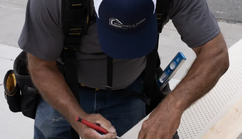 A Windows and Siding Reno installer in a logo cap bends over a perforated soffit panel, holding a red pencil to its edge
