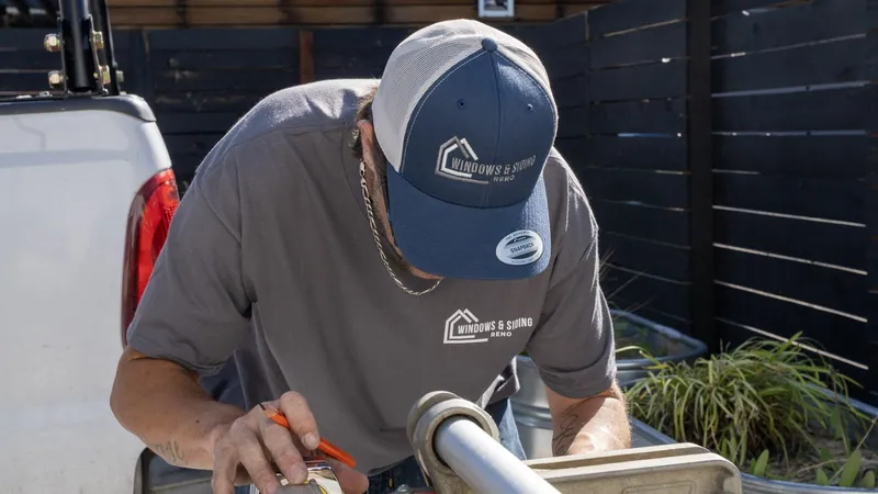 A Windows and Siding Reno installer in a logo cap holds a tape measure and pencil at a siding brake by a white pickup
