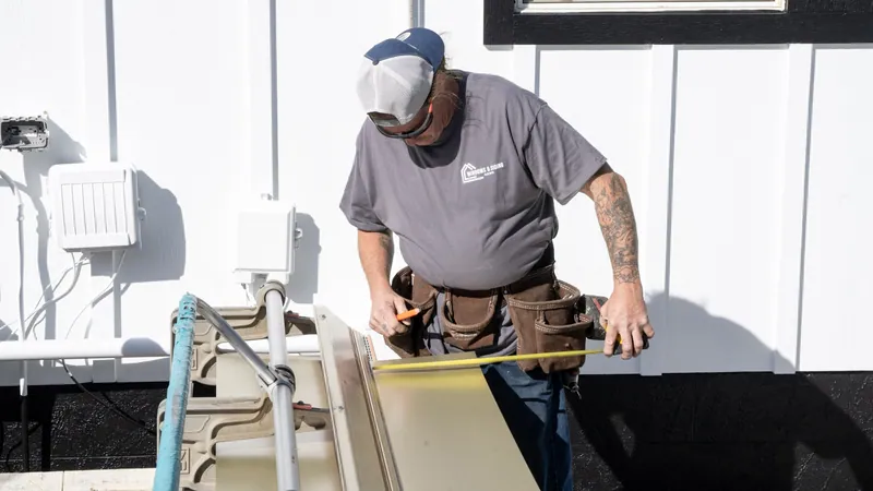 A Windows and Siding Reno installer measures trim coil on a brake beside white board and batten siding with a black base