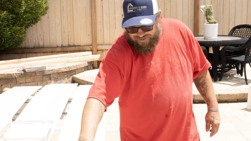 A Windows and Siding Reno crew member in a logo cap and red tee stands over long white panels and a paint tray on a patio