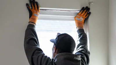 An installer seen from behind presses both gloved hands to the head of a tall new white window from inside a room