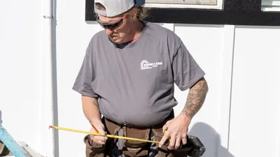 A Windows and Siding Reno installer in safety glasses and a leather tool belt stretches out a yellow tape measure
