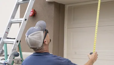 An installer extends a yellow tape measure straight up in front of a garage door, with two ladders beside him