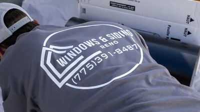 A Windows and Siding Reno installer unboxes a roll of black trim coil on a drop cloth, the logo across his shirt back