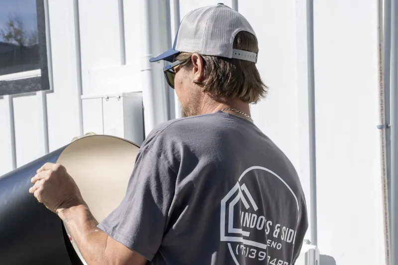 A Windows and Siding Reno installer unrolls black trim coil beside a white board and batten wall with utility boxes