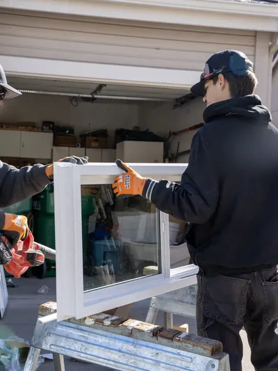 Two installers in work gloves lifting a white vinyl slider window unit off a padded stand in front of an open garage