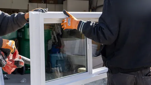 Close view of two installers in work gloves holding up a white vinyl window unit, with an open garage behind them