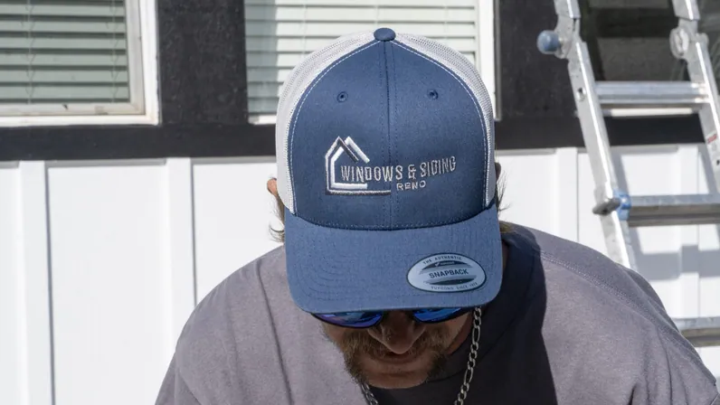 A Windows and Siding Reno crew member looks down in a logo cap, white framed windows in a dark wall and a ladder behind