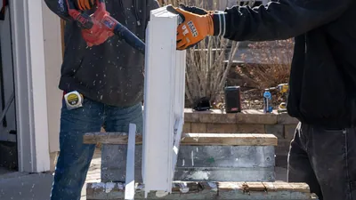 Two installers stand a new white vinyl window frame on a sawhorse and blow it clean in a sunny driveway