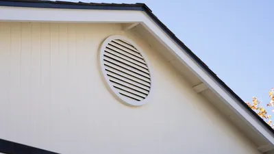 A round louvered gable vent set into cream vertical grooved panel siding under white fascia and a shingle roof edge