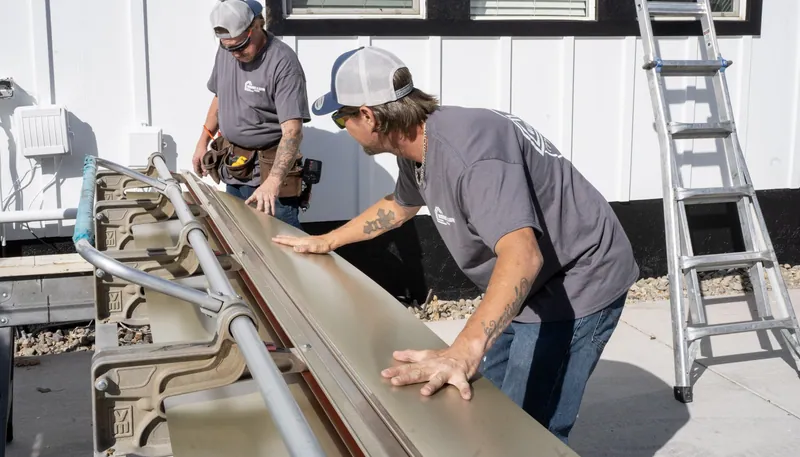 Two Windows and Siding Reno installers set tan trim coil in a brake beside white board and batten siding with a black base