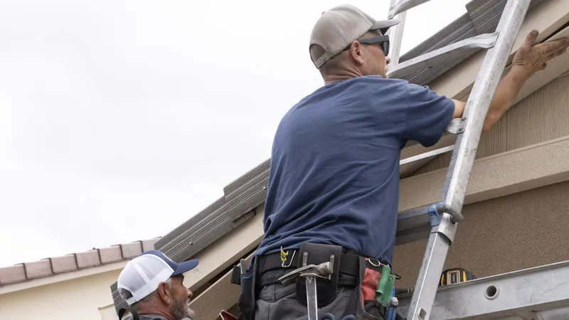 Two installers work at a tile roof edge above a stucco wall, one reaching up from an aluminum extension ladder