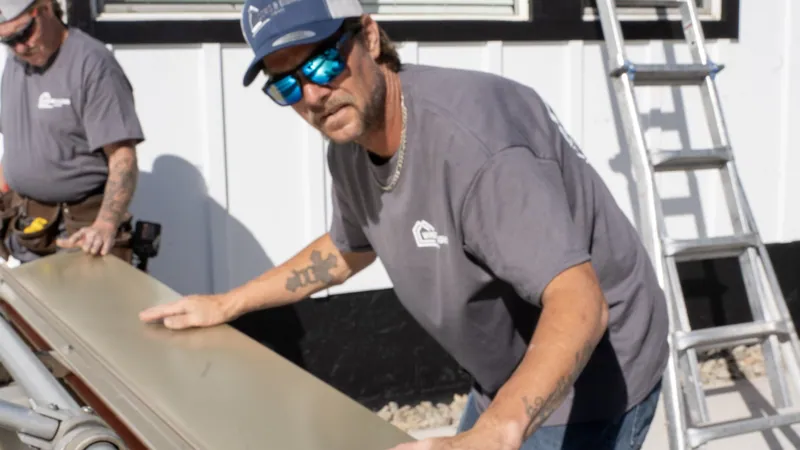 Two installers in logo tees position tan trim coil on a sheet metal brake beside white board and batten siding