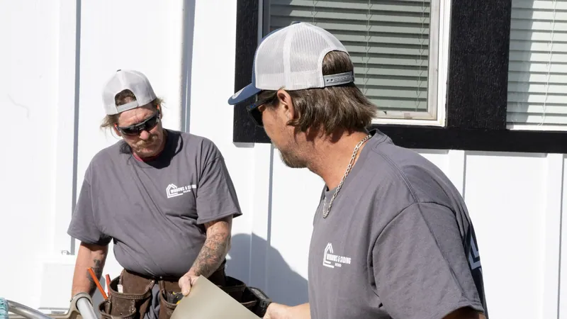 Two Windows and Siding Reno installers in logo tees talk over a tan trim sheet beside a white window with black trim