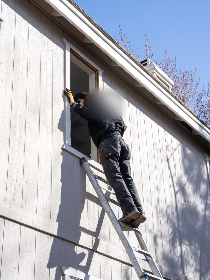 Installer on an extension ladder working the jamb of an upper-storey window opening